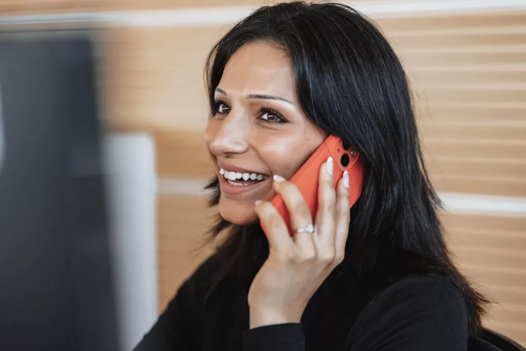 Eine lächelnde Frau mit dunklen Haaren telefoniert mit einem roten Smartphone. Sie sitzt an einem Schreibtisch und wirkt freundlich und konzentriert.