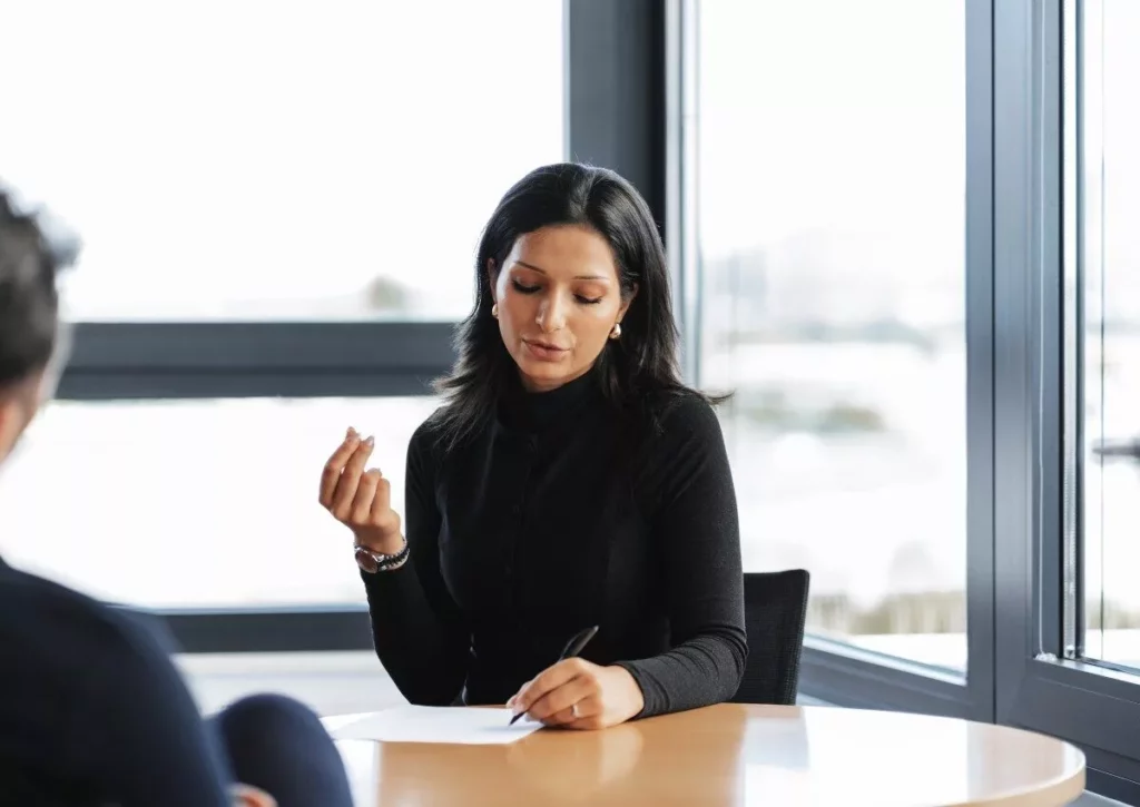 Eine Frau sitzt an einem Tisch in einem modernen Büro, schreibt auf ein Blatt Papier und spricht mit einer anderen Person. Sie wirkt konzentriert und professionell.
